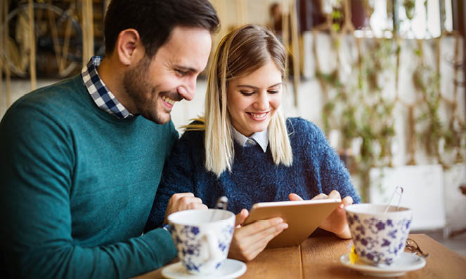 happy-couple-in-sweaters-looking-at-tablet-with-floral-tea-cups