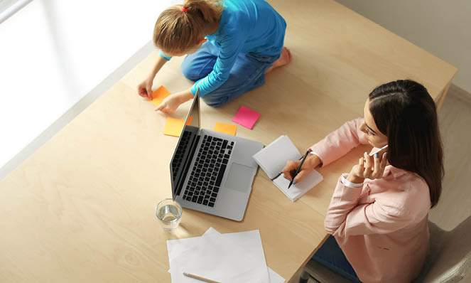 mother-working-desk-looking-at-laptop-daughter-sitting-with-sticky-notes