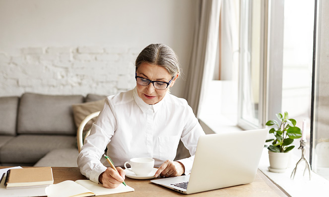 older-woman-sitting-at-home-writing-on-notebook-silver-laptop-open