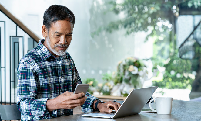 oldern-man-flannel-shirt-grey-beard-looking-at-phone-and-laptop-sitting-by-window